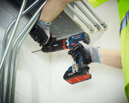 Worker using Bosch cordless drill to drill into a white wall in an industrial setting; wearing gloves and high-visibility shirt with conduit pipes visible overhead.