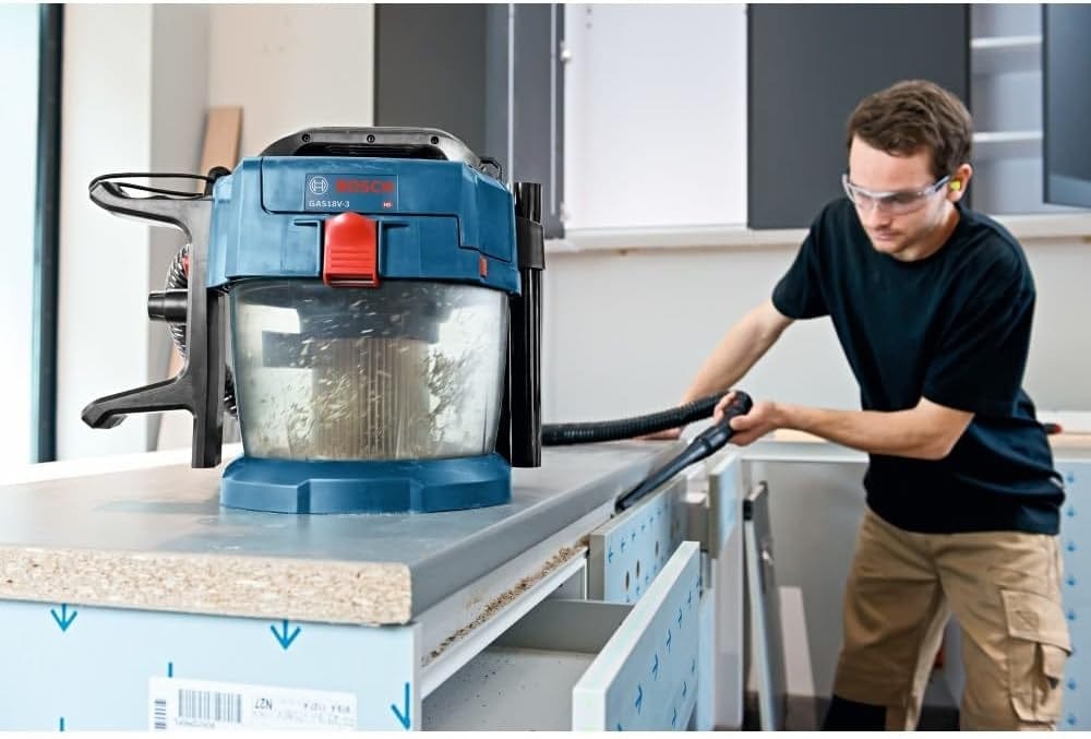 Worker using Bosch GAS18V-10L cordless vacuum to clean a laminated countertop in a workshop, wearing safety goggles and work attire.