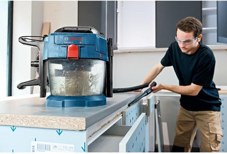 Worker using Bosch GAS18V-10L cordless vacuum to clean a laminated countertop in a workshop, wearing safety goggles and work attire.