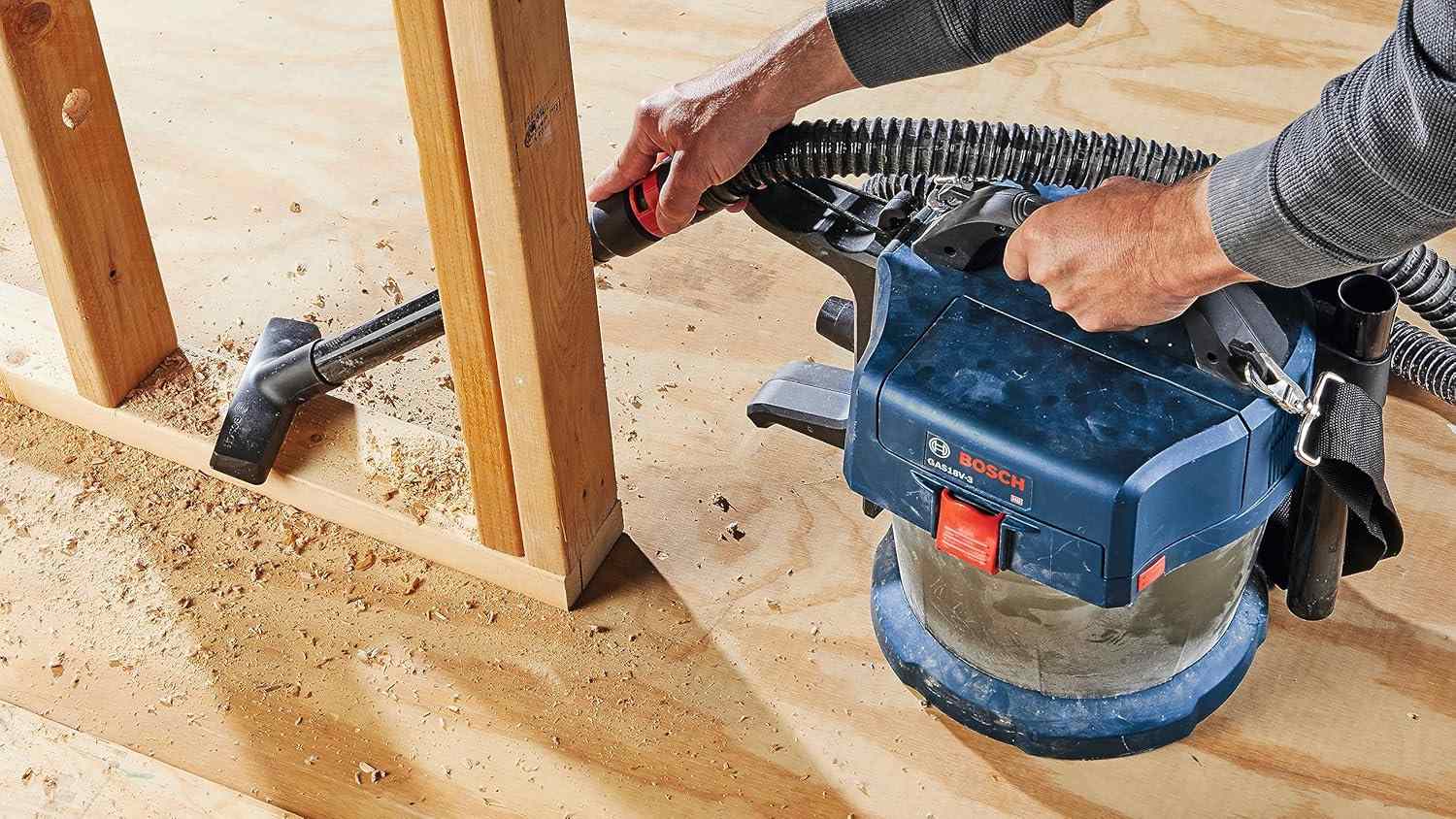Person using Bosch cordless wet/dry vacuum to clean sawdust from a wooden floor near a framed wall structure at a construction site.