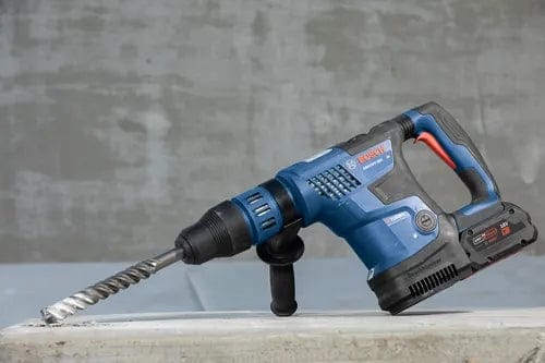 Bosch rotary hammer drill with spiral bit partially embedded in concrete, shown mid-operation with side handle for control and stability.