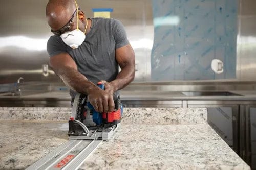 Person using Bosch track saw with guide rail to cut stone countertop, wearing safety gear in a modern kitchen or workshop setting