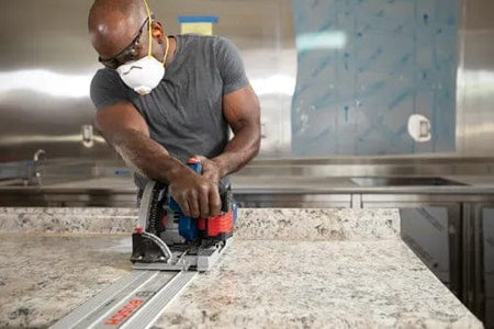 Person using Bosch track saw with guide rail to cut stone countertop, wearing safety gear in a modern kitchen or workshop setting