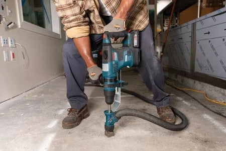 Person using a Bosch hammer drill with dust extraction to drill into a concrete floor, wearing gloves and work boots in a construction setting.