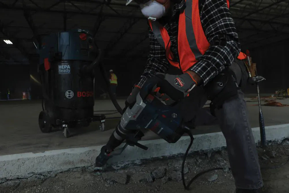 A construction worker in high-visibility safety gear using a Bosch SDS-Max combination hammer for concrete floor demolition while connected to a HEPA-ready dust extraction vacuum system.