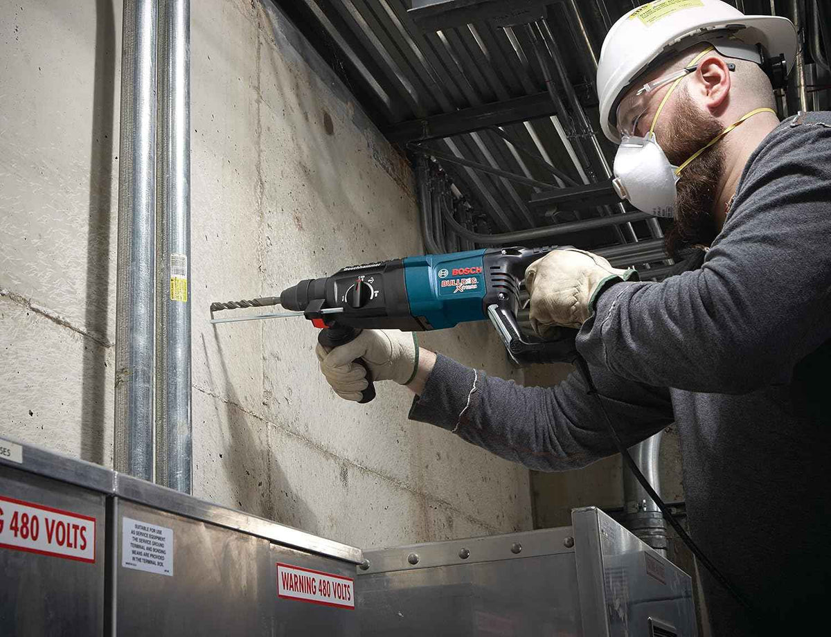 A construction worker wearing a white hard hat and safety glasses drilling into a concrete wall in an industrial setting with exposed conduit and safety signage.