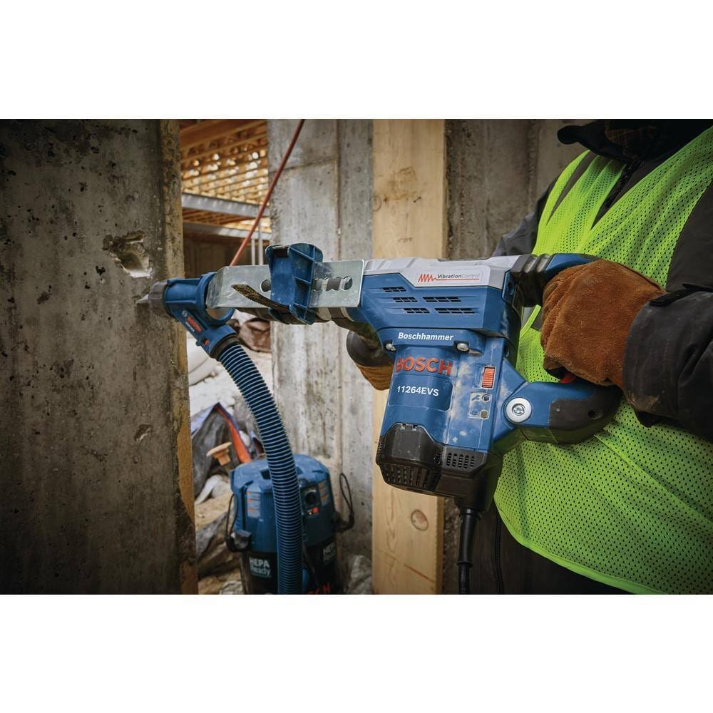 A construction worker wearing a high-visibility vest drilling horizontally into a concrete wall, using the combination hammer connected to a dust extraction attachment and vacuum hose.