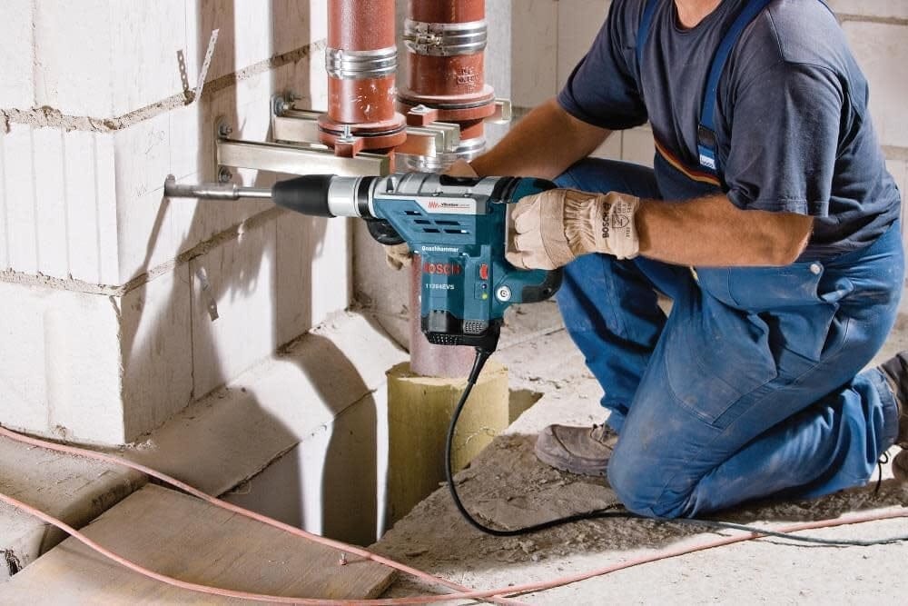A construction worker kneeling to drill into a concrete foundation wall near plumbing pipes using the combination hammer.