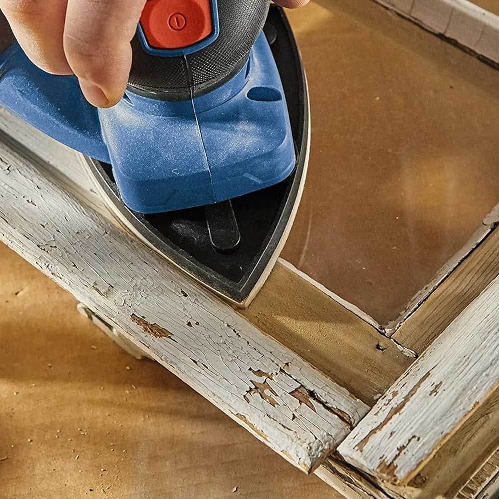 Person using Bosch detail sander with pointed pad to smooth chipped paint on wooden window frame; workshop setting visible.