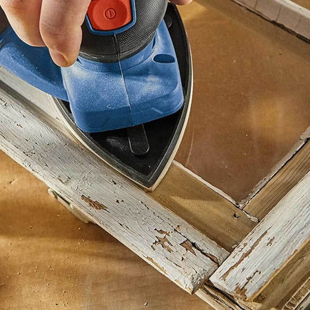 Person using Bosch detail sander with pointed pad to smooth chipped paint on wooden window frame; workshop setting visible.