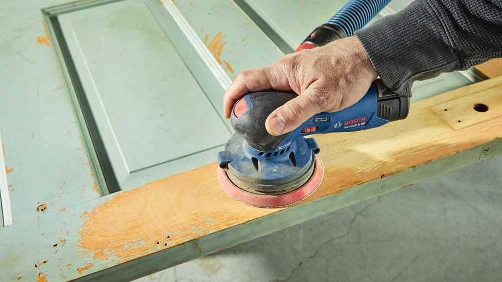 Person using Bosch electric sander with vacuum hose to remove paint from wooden door surface in restoration task.