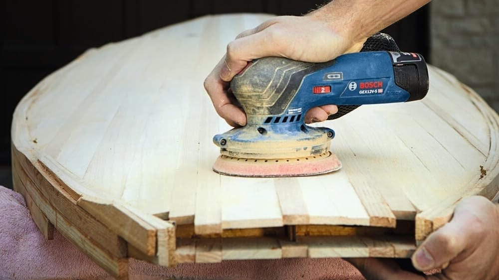 Person using Bosch GEX 125-1 AE orbital sander to smooth light-colored wood surface; tool held with one hand.