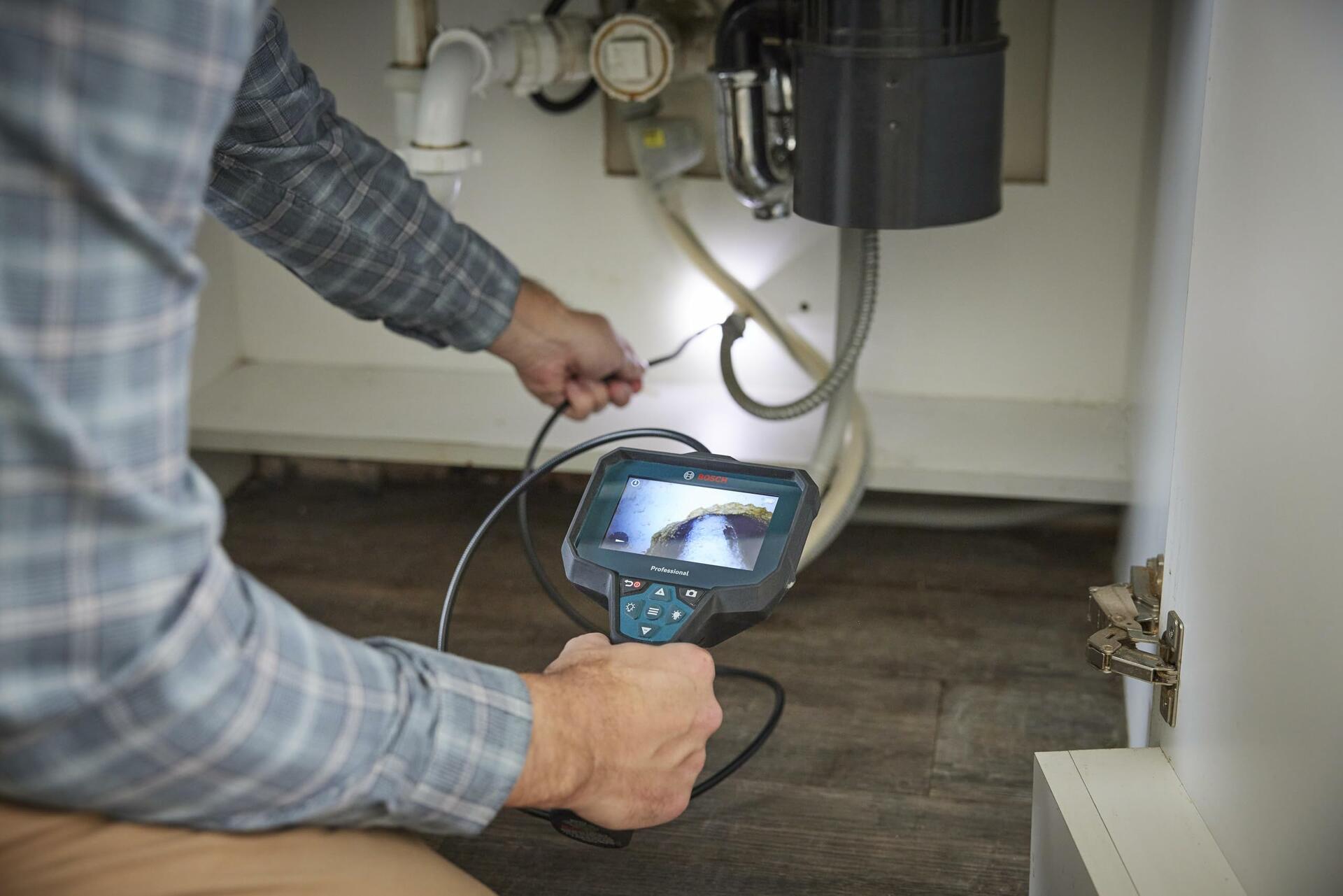 A person kneels to inspect plumbing under a cabinet, holding the Bosch inspection camera to view the hard-to-reach area behind a garbage disposal.