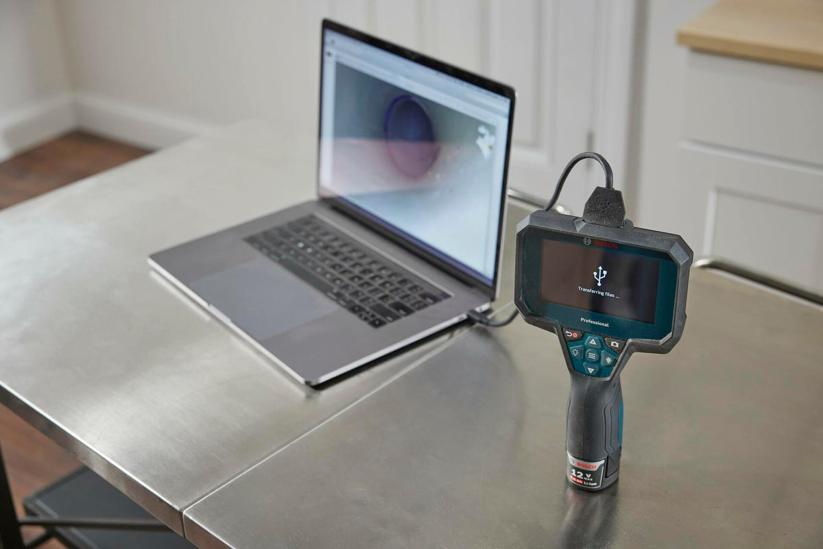 The Bosch GIC4-23C inspection camera on a metal workbench, connected via USB-C cable to a laptop, demonstrating the file transfer feature with an image displayed on both screens.