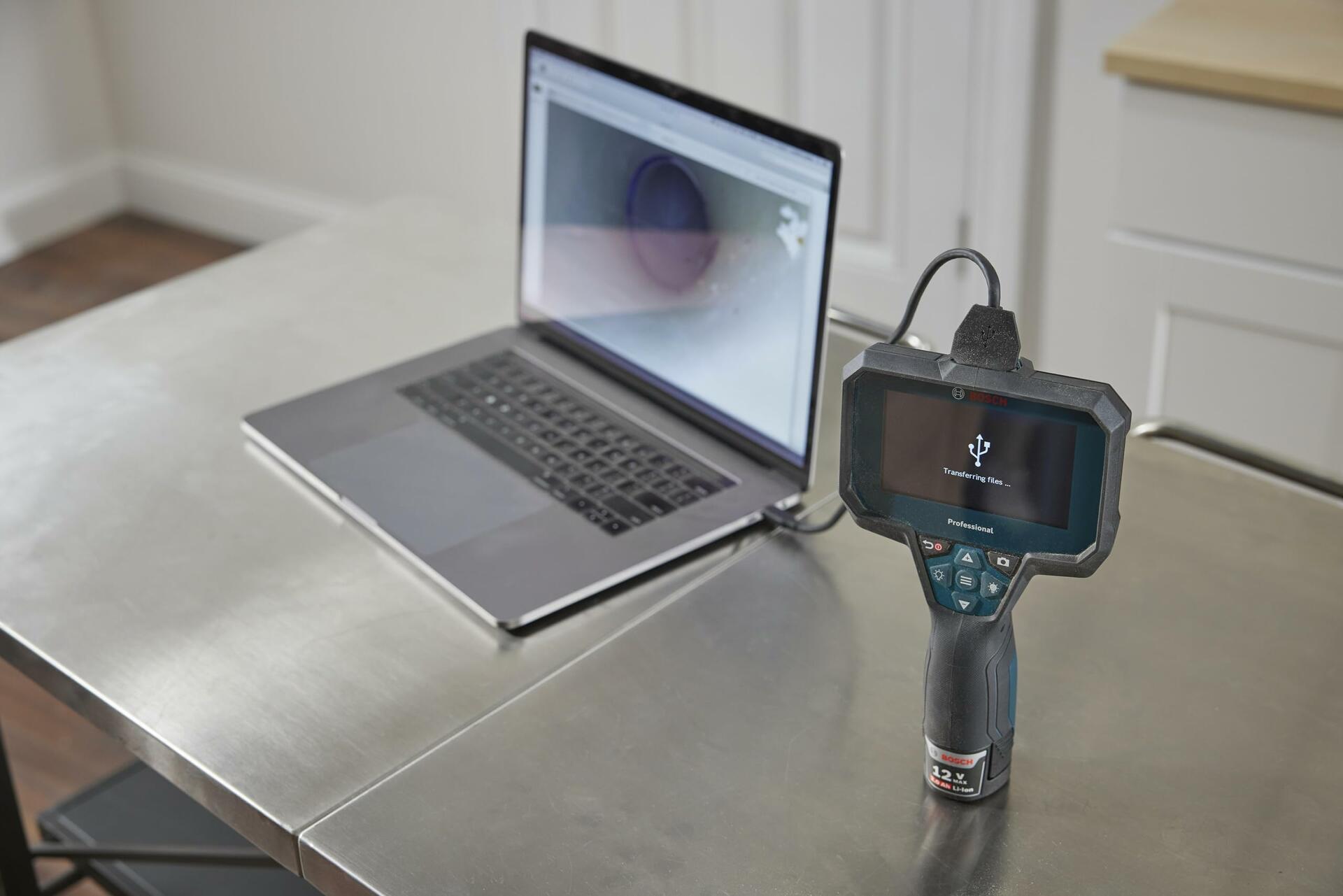 The Bosch GIC4-23C inspection camera on a metal workbench, connected via USB-C cable to a laptop, demonstrating the file transfer feature with an image displayed on both screens.