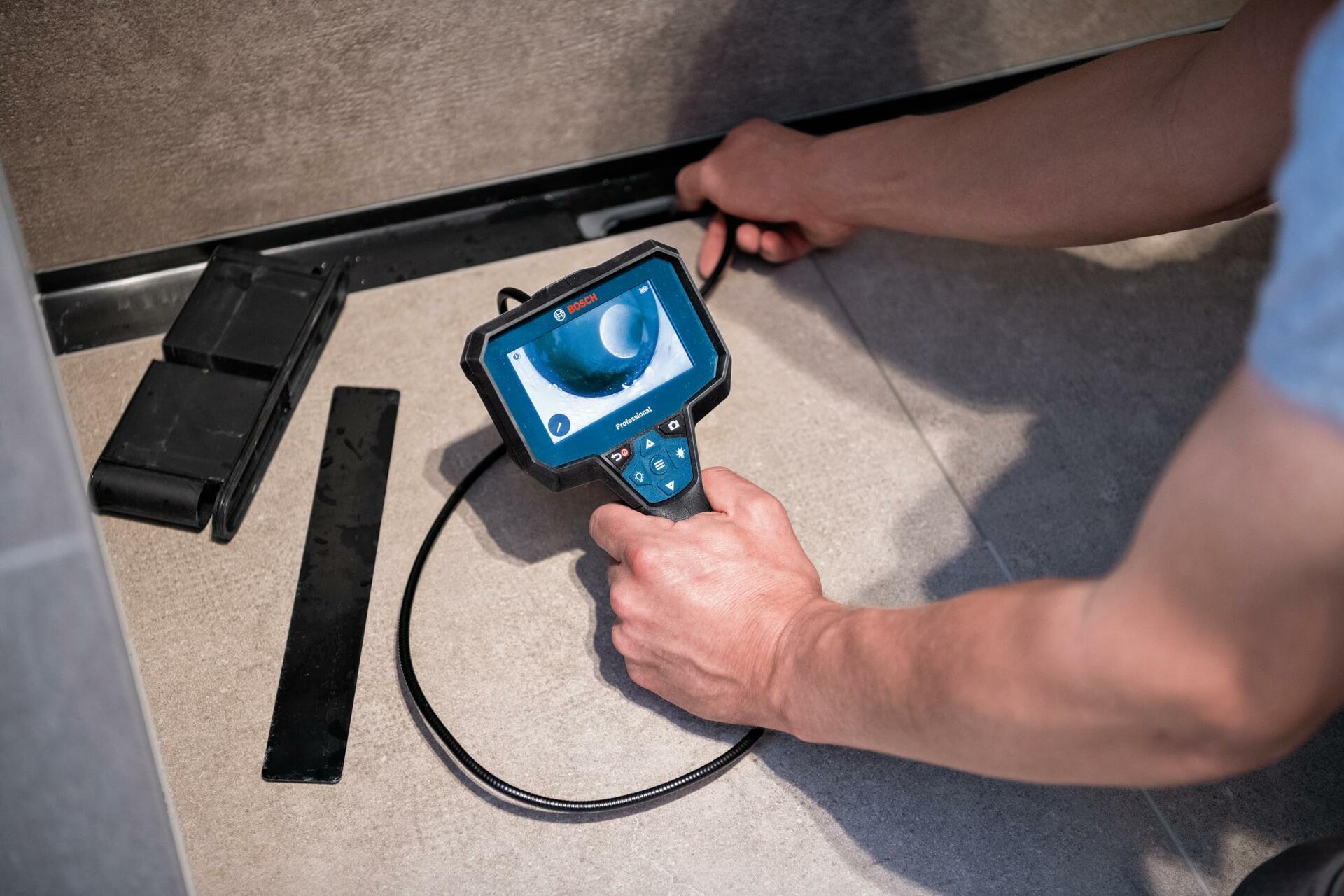 A person uses the Bosch inspection camera to look inside a floor drain, holding the display in one hand while guiding the flexible cable with the other.
