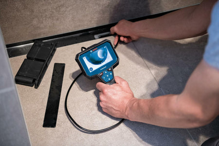 A person uses the Bosch inspection camera to look inside a floor drain, holding the display in one hand while guiding the flexible cable with the other.