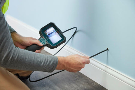 A worker feeds the flexible camera cable of the Bosch inspection camera into a small hole in a wall near the baseboard to inspect the internal wall cavity.