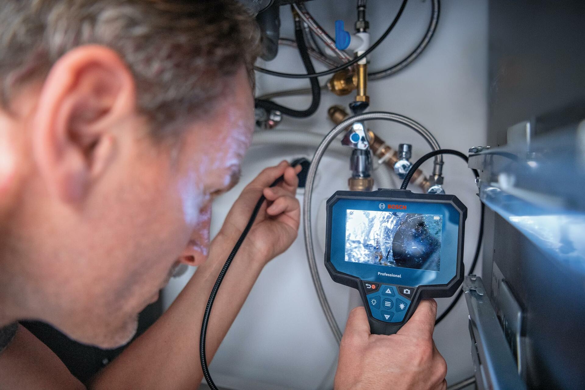 A plumber holds the Bosch inspection camera, inserting the camera cable to get a clear view of the complex pipes and valves of a water system on the large display.