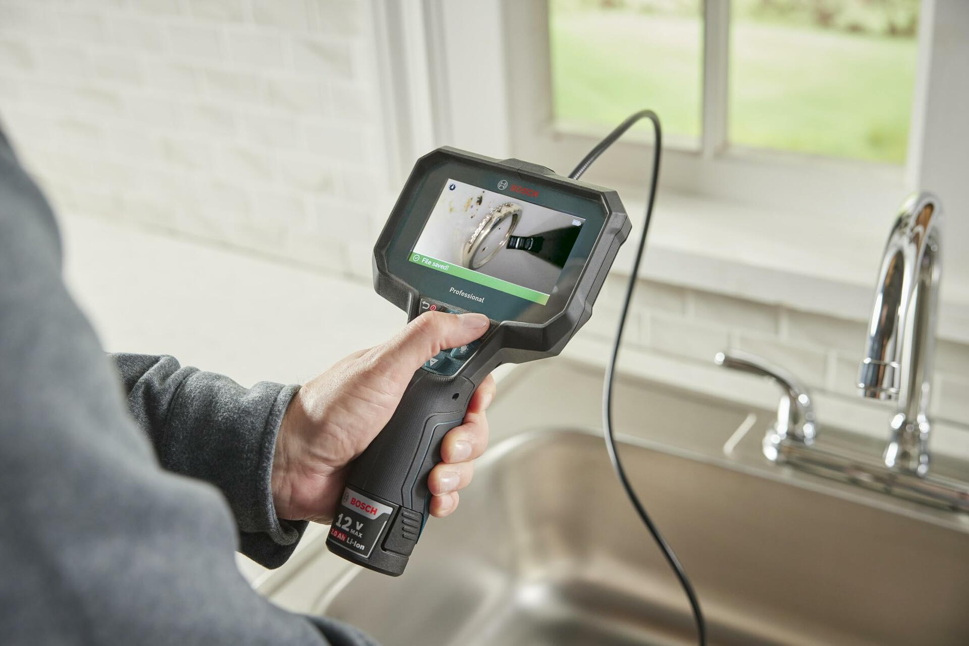 A person holds the Bosch inspection camera over a kitchen sink, and the screen displays a "File saved" confirmation after capturing an image of a ring lost in the drain.