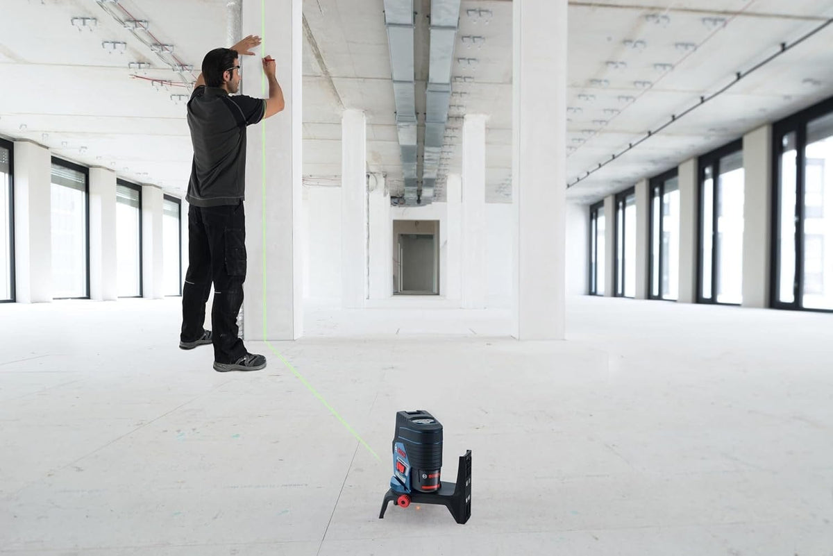 Worker uses Bosch green-beam laser level on a tripod to align vertical and horizontal lines on a column inside a large unfinished commercial building