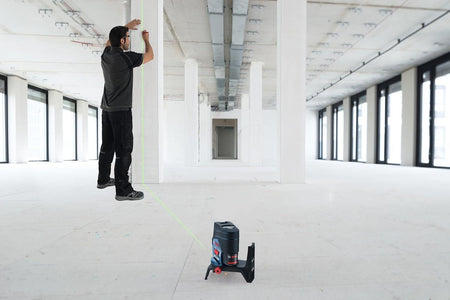 Worker uses Bosch green-beam laser level on a tripod to align vertical and horizontal lines on a column inside a large unfinished commercial building
