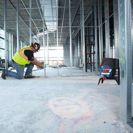 Construction worker kneels on concrete floor using Bosch laser level with red beam to align metal framing studs inside a partially built interior space.