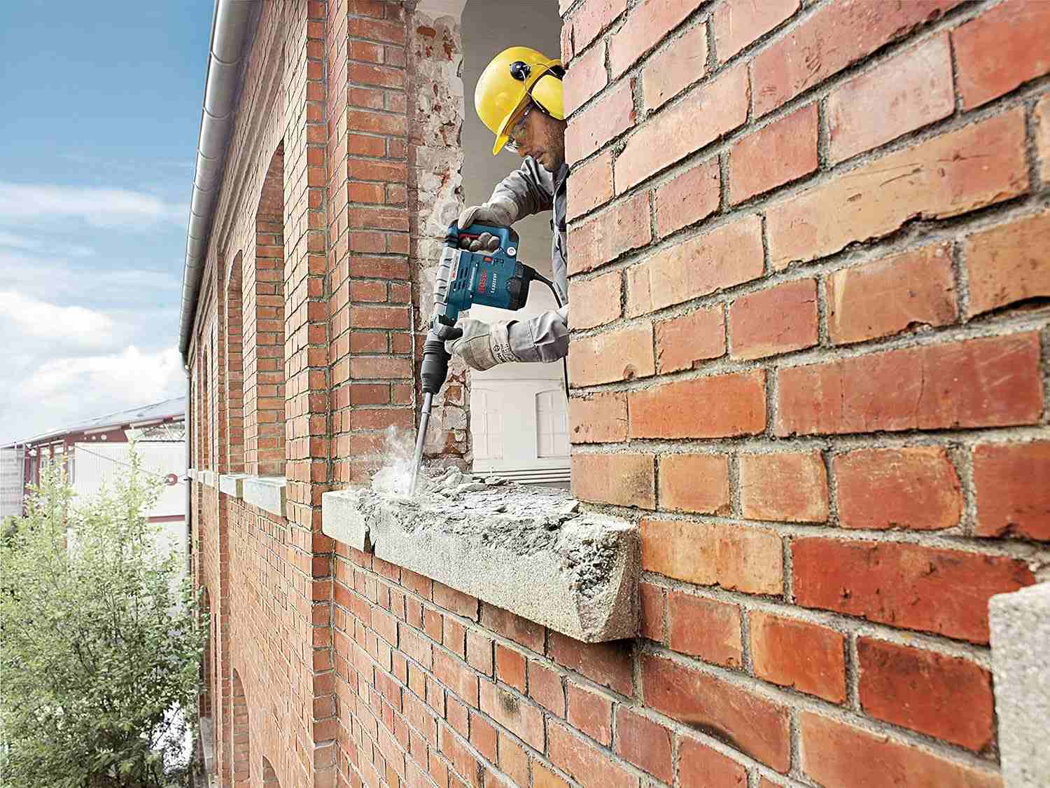 An action shot of a worker in safety gear using the Bosch 11321EVS demolition hammer to chisel a concrete window sill on a brick building.