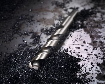 Macro shot of a cobalt drill bit lying on a metal surface surrounded by dark metal drilling chips.