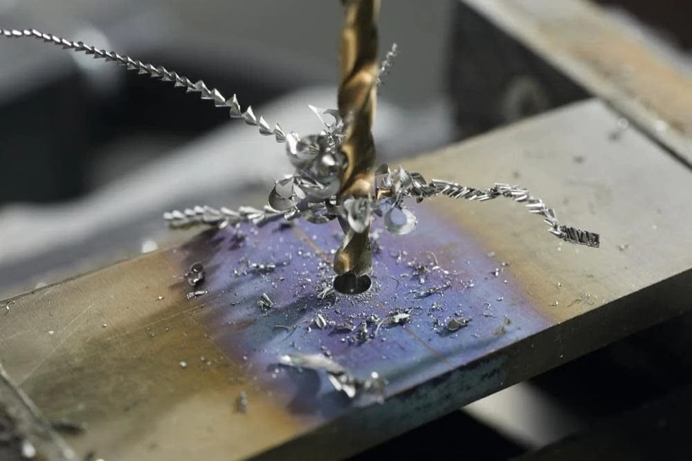 Extreme close-up of a cobalt bit drilling into metal, producing long, spiral metal shavings with heat discoloration visible on the workpiece.