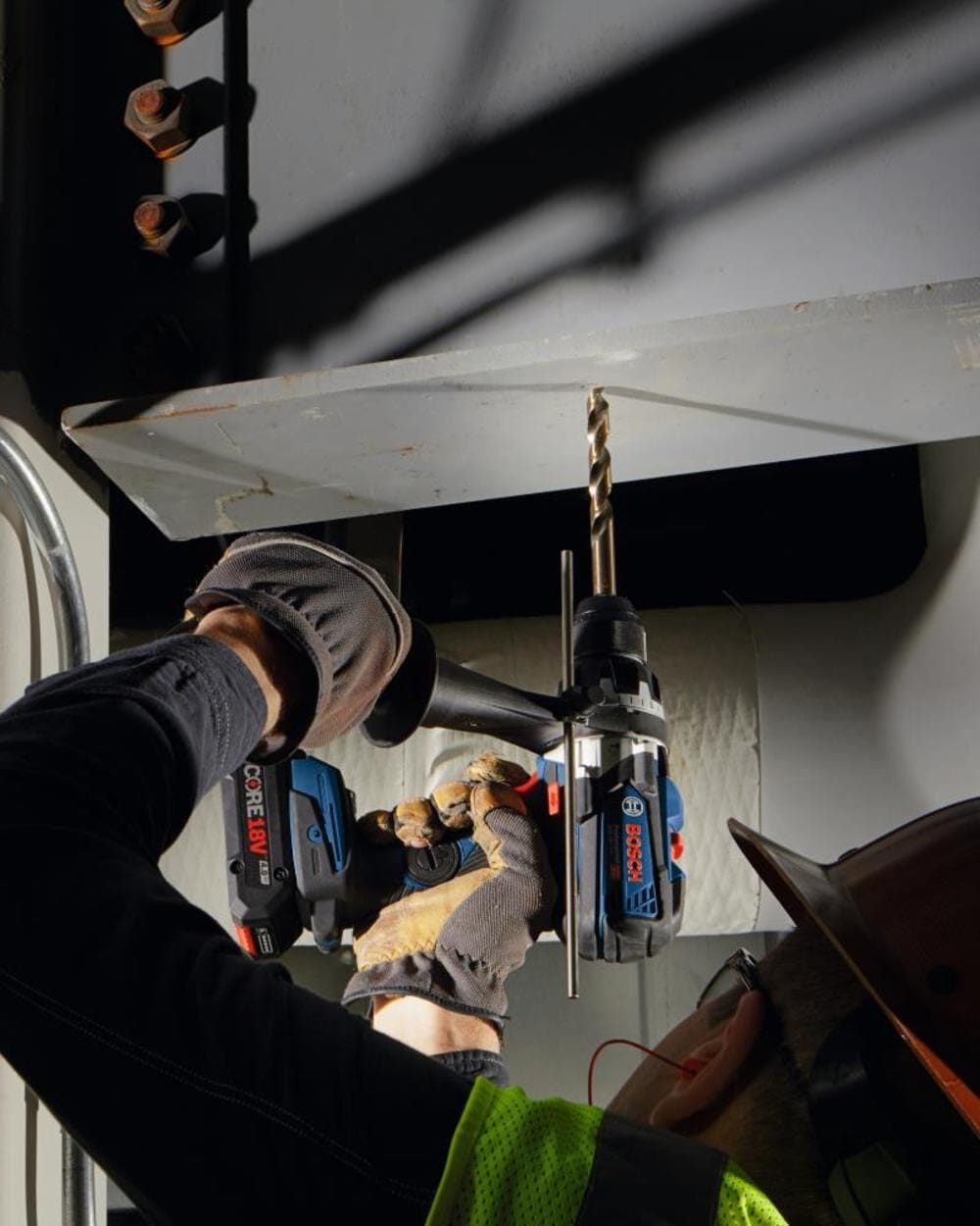 A worker drilling upwards into a steel I-beam using a Bosch cordless drill and a gold-tone cobalt drill bit.