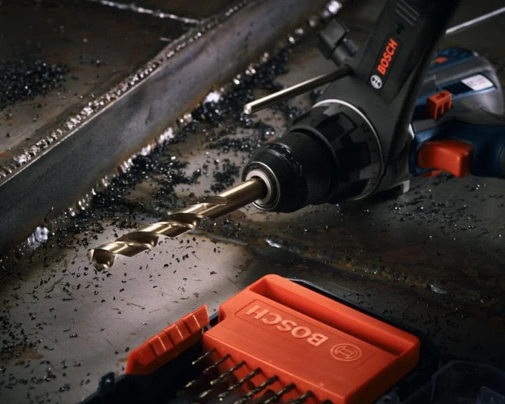 High-angle view of a drill with a cobalt bit resting on a metal surface covered in shavings next to a completed hole.