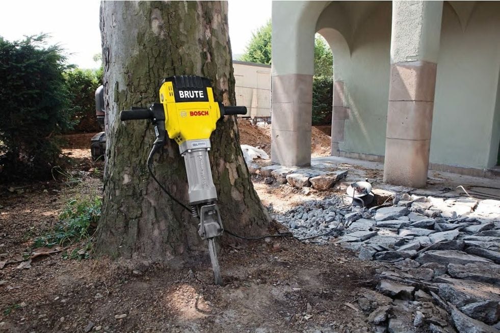 Bosch Brute jackhammer mounted vertically against tree trunk at construction site with broken concrete and debris.