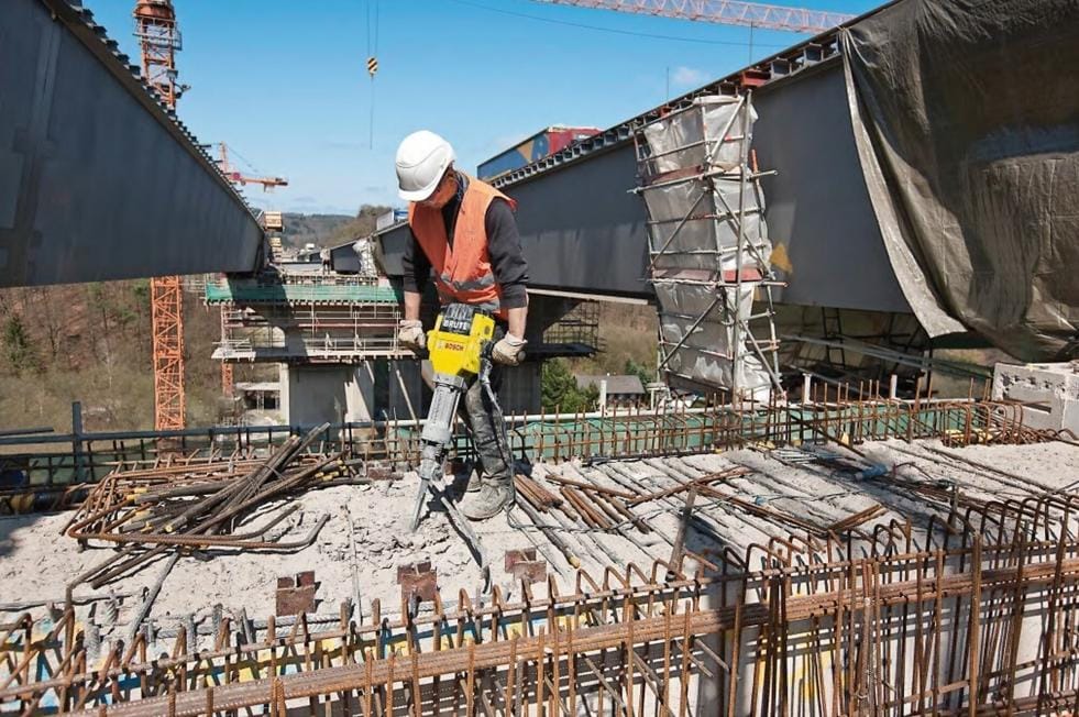 Worker operating jackhammer on bridge construction site; steel beams, scaffolding, and cranes visible in background.