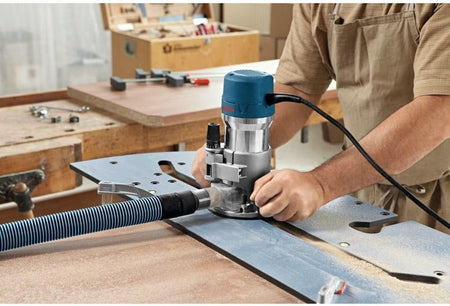 Professional action shot of a woodworker using the Bosch 1617EVS router on a workbench with a dust extraction hose to mill a precise groove along a large wooden board.