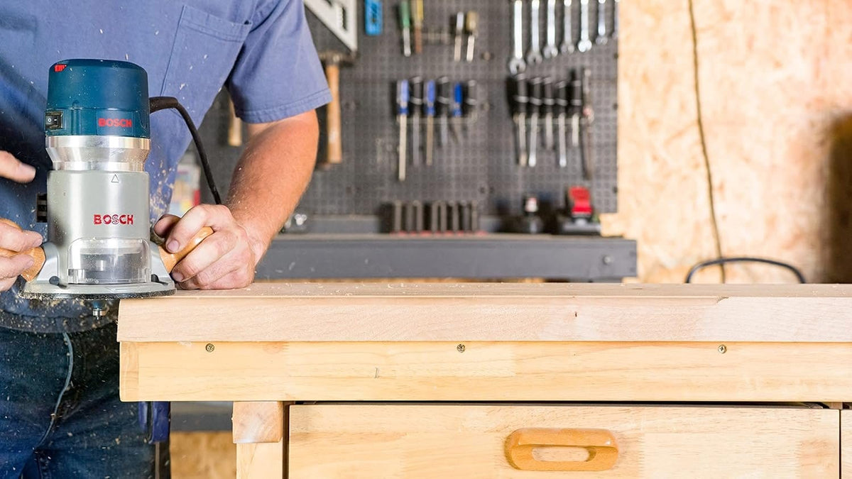 Action shot of a technician using the Bosch 2.25 HP corded router to trim the edge of a wood panel, illustrating the tool's stability and power for consistent edge-forming.