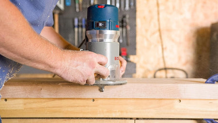 Side action view of the Bosch fixed-base router in operation on a workbench, highlighting the dust-sealed power switch and smooth glide across a hardwood surface.