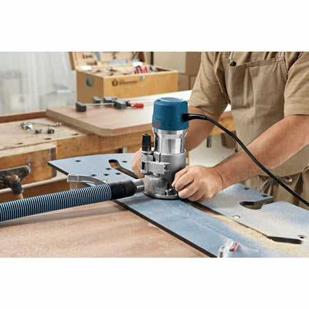 Person using Bosch router connected to vacuum hose for dust collection. Router guided along metal jig on workbench. Workshop setting includes apron, clamps, screws, and tools.