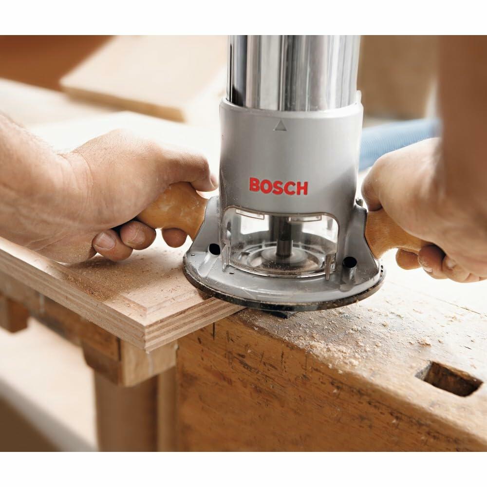 Close-up of Bosch router shaping the edge of a wooden board. User grips hardwood handles while guiding the tool along the surface. Workshop background with other wood pieces visible.