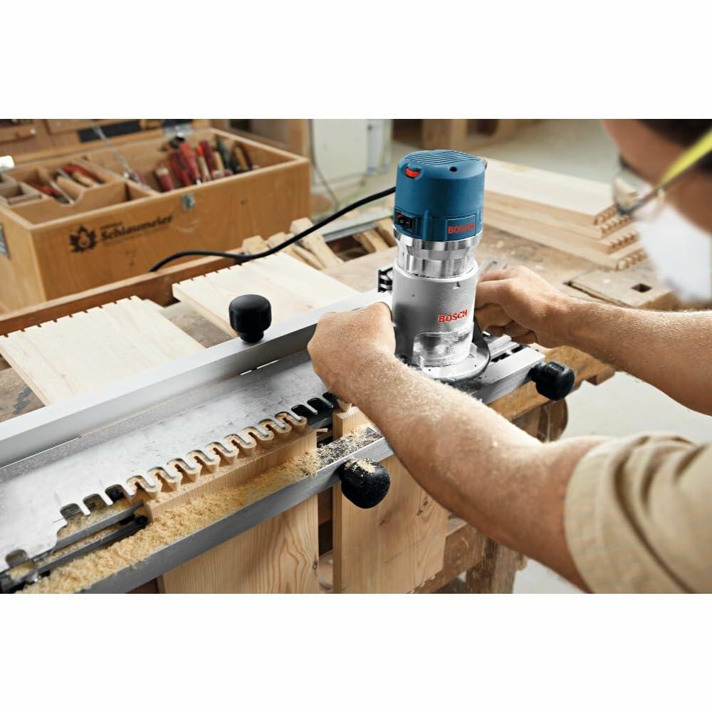 Person using Bosch router with dovetail jig to cut interlocking joints into wooden boards. Workbench setup includes hand tools and a wooden toolbox in a woodworking environment.