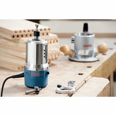 Two Bosch routers on a woodworking bench surrounded by dovetail joint samples. Foreground router is compact with accessories nearby; background router has hardwood handles.