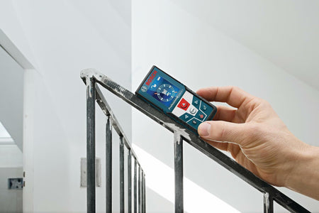 Close-up of a hand holding the Bosch GLM50C on a staircase railing, using the inclinometer function to measure and display the 36.4 angle of the slope