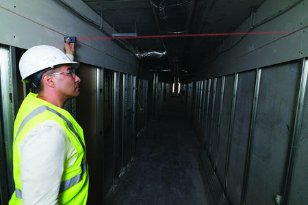 Construction worker aiming the Bosch GLM50C laser measure down a long industrial corridor framed with metal studs, demonstrating long-range measurement capability