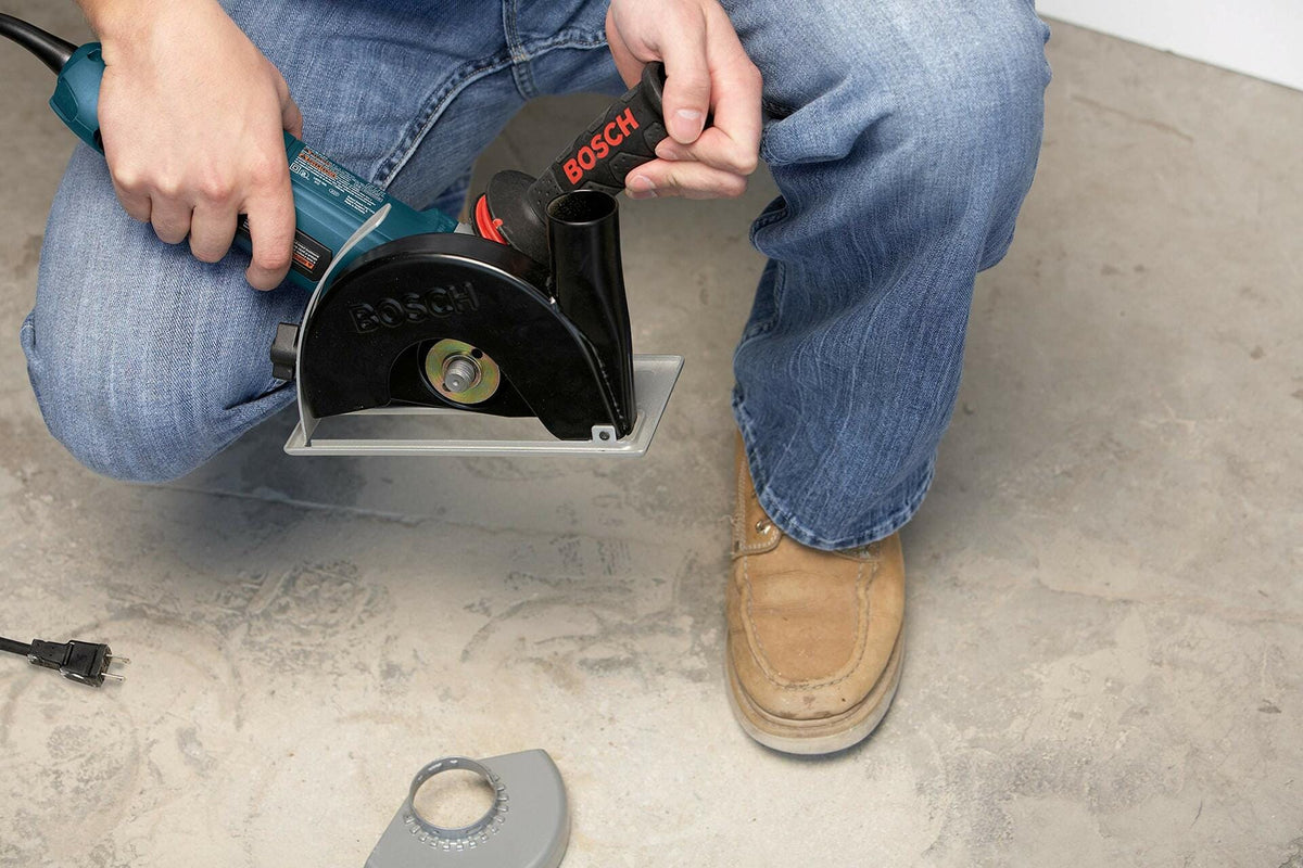 Person using a Bosch wall chaser on a concrete floor with vacuum port attached. Tool features a circular blade and protective casing for dust control. Scene shows active use in a construction setting with visible power cord and accessory nearby.