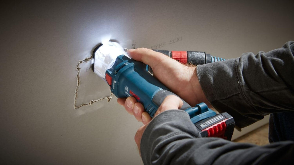 A person uses the Bosch GCU18V-30 brushless cut-out tool to cut a hole in a sheet of drywall, with the tool's LED light illuminating the cut line.