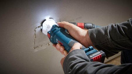 A person uses the Bosch GCU18V-30 brushless cut-out tool to cut a hole in a sheet of drywall, with the tool's LED light illuminating the cut line.