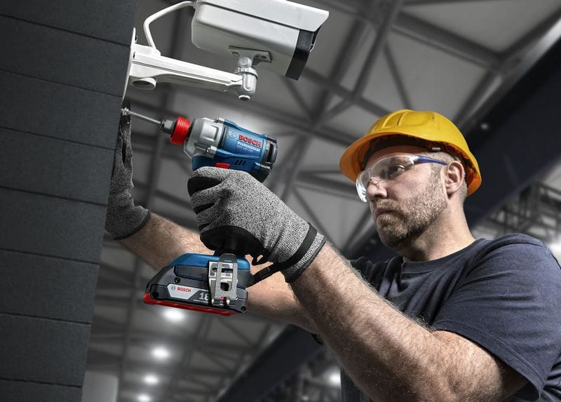 An electrician wearing a yellow safety hard hat and transparent protective goggles uses a long screwdriver bit inside the Bosch hybrid 1/4-inch hex chuck to mount an outdoor commercial security camera housing onto a dark tile wall structure.