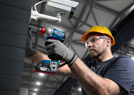 An electrician wearing a yellow safety hard hat and transparent protective goggles uses a long screwdriver bit inside the Bosch hybrid 1/4-inch hex chuck to mount an outdoor commercial security camera housing onto a dark tile wall structure.