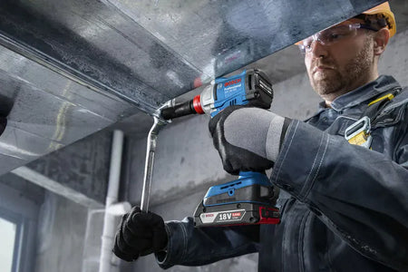 An HVAC installation specialist wearing protective safety glasses, thick gloves, and a tool harness uses the Bosch combination impact driver wrench tool to secure overlapping joints on an industrial metal ventilation duct system. The operator uses an open-end hand wrench alongside the high-torque cordless tool to lock a bracket bolt into position.