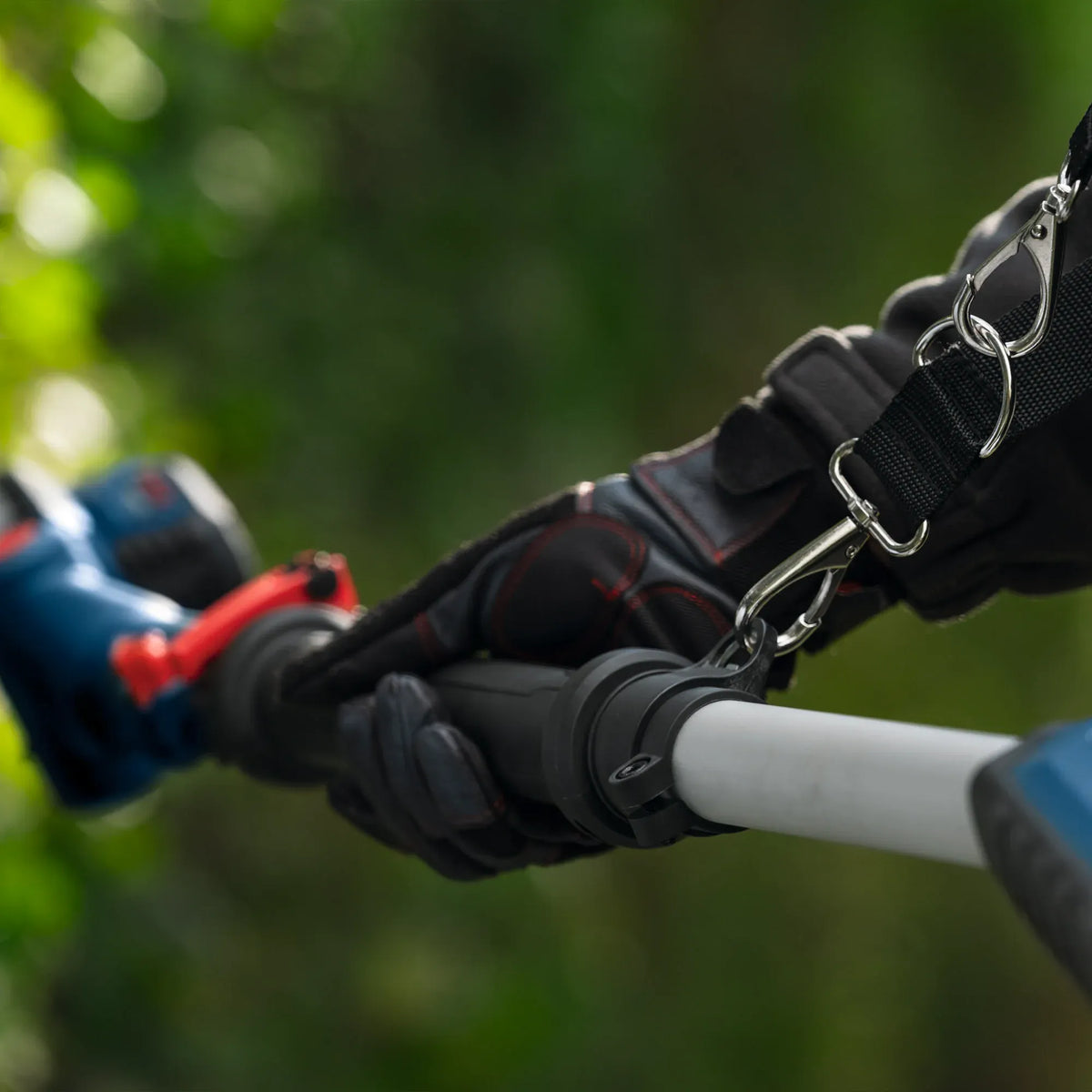 A close-up macro view displays a gardener's gloved hand holding the main pole handle grip of the Bosch hedge trimmer. The image focuses on the quick-release metal clip attaching the heavy-duty supportive shoulder safety strap to the trimmer's alignment ring.